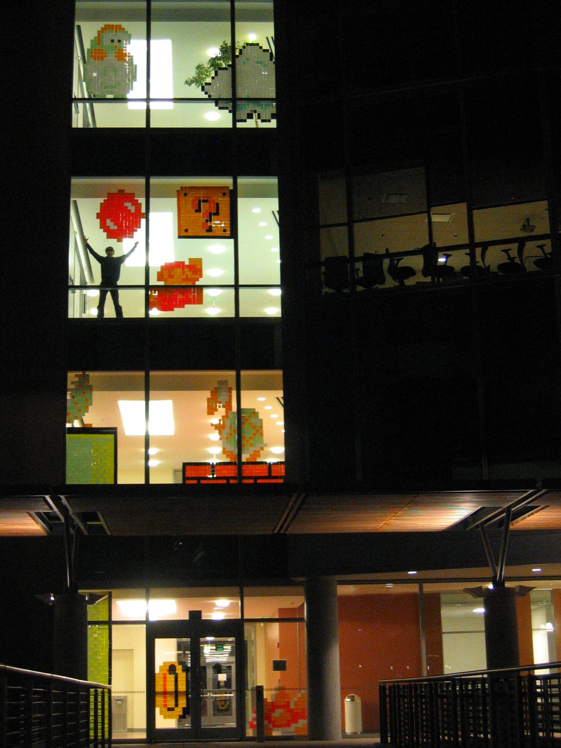 The finished installation at night: four floors of windows filled with Post-it sprites, and a figure with arms raised on the third floor